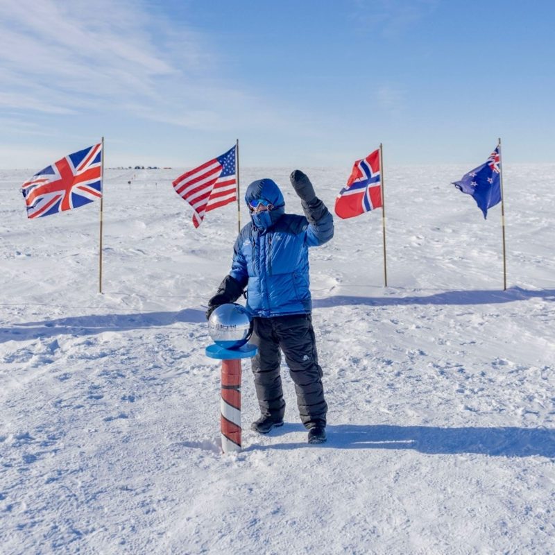 Ashley Scott Robertson in a blue down jacket at the South Pole marker, waving, with the UK, US, Norway and New Zealand flags behind