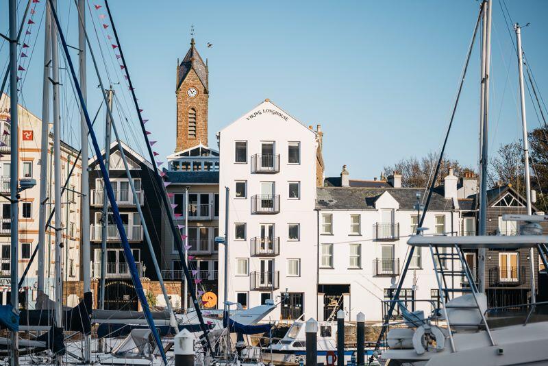 The white Viking Longhouse building beside the yachts and pontoons of Peel Marina, with the clock tower of the old church behind